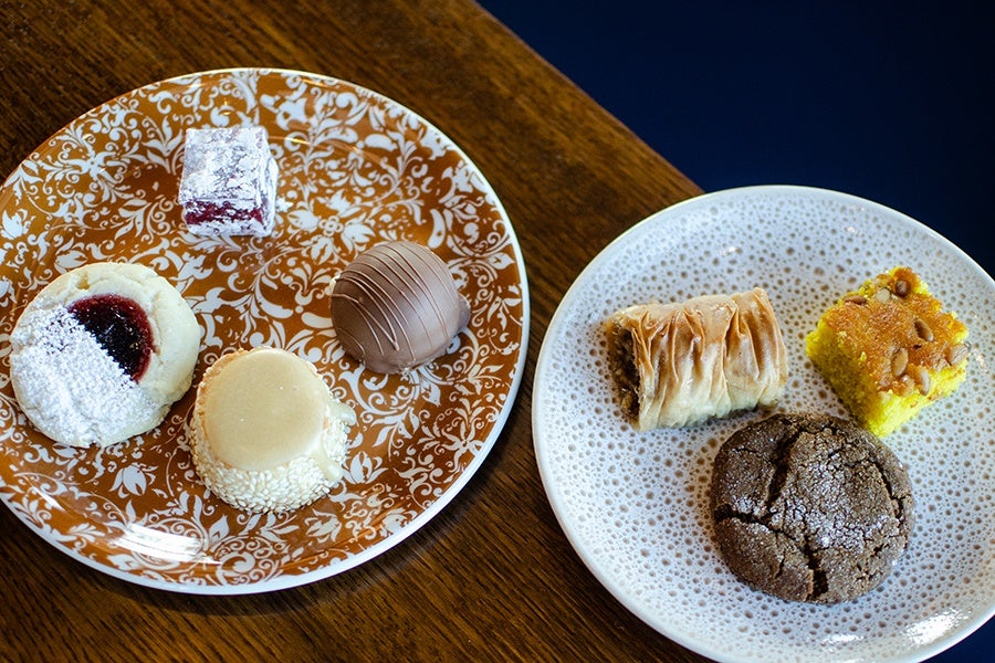 Two decorative plates on a wooden surface, each with an assortment of desserts. The left plate has a powdered sugar-dusted thumbprint cookie with jam, a sesame-coated round treat, a chocolate truffle, and a square red jelly candy dusted with powdered sugar. The right plate holds a piece of baklava, a yellow square cake topped with nuts, and a cracked chocolate cookie dusted with sugar.