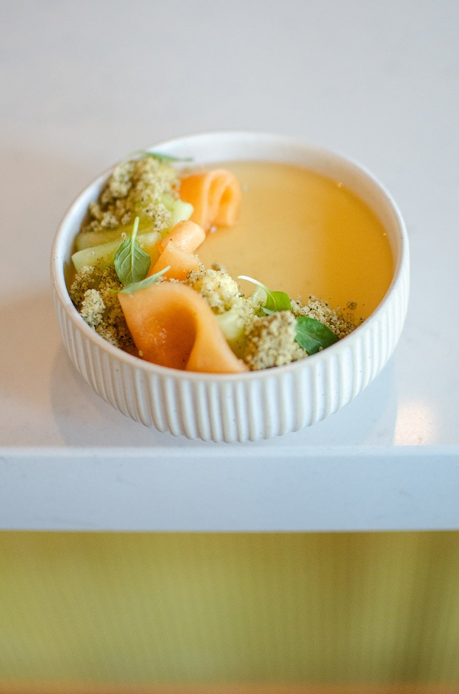 A white ribbed bowl containing a light yellow dessert garnished with thin orange melon ribbons, green leaves, and a crumbly topping. The bowl is placed on a white surface with a yellow background below.