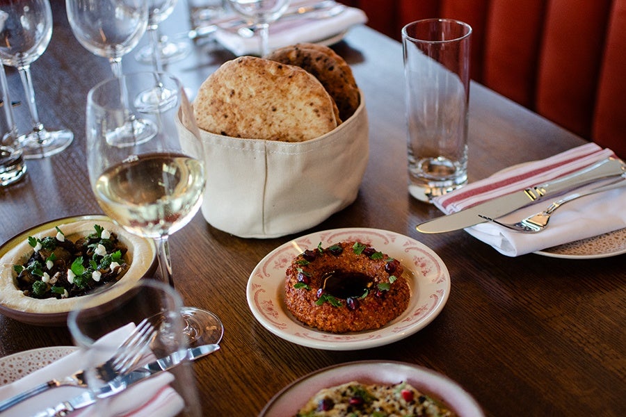 A wooden table set with a basket of pita bread, a plate of red dip garnished with herbs and pomegranate seeds, a bowl of hummus topped with herbs and pine nuts, a glass of white wine, an empty tall glass, and neatly folded napkins with silverware. Several empty wine glasses are also visible in the background.