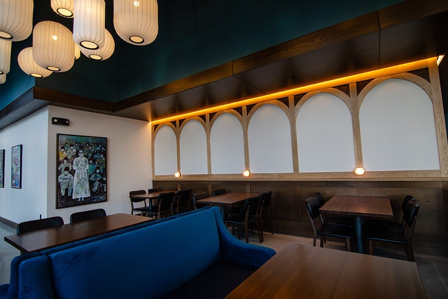 A modern dining area with wooden tables and black chairs, featuring a blue upholstered bench in the foreground. The wall has decorative wooden arches with white panels and small round lights beneath a strip of warm yellow lighting. Several large, white, cylindrical hanging lanterns illuminate the space, and there are framed artworks on the adjacent wall.