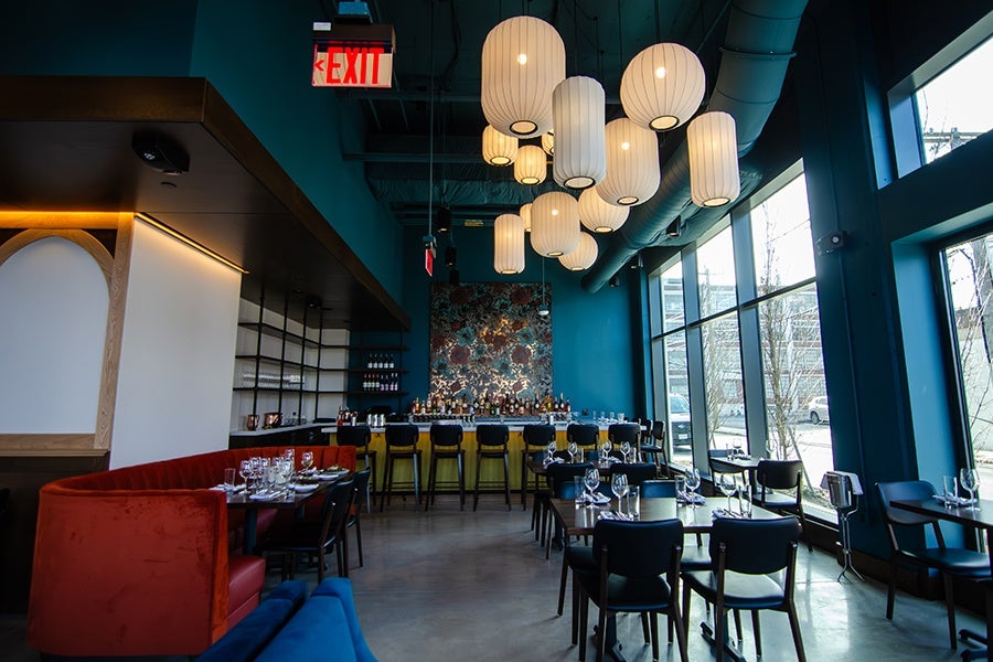 Modern restaurant interior with teal walls, large windows, and a bar area featuring a floral patterned backdrop. The seating includes a curved red velvet booth, black chairs around dark wooden tables, and yellow bar stools. Multiple white, cylindrical pendant lights hang from the ceiling, and an illuminated exit sign is visible above the bar. Tables are set with wine glasses and cutlery.