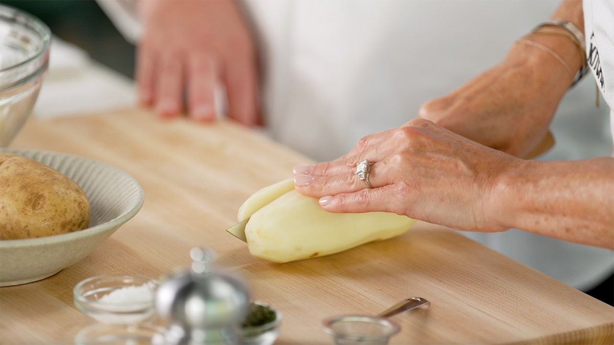 A chef slices a potato longways on a cutting board in a kitchen.
