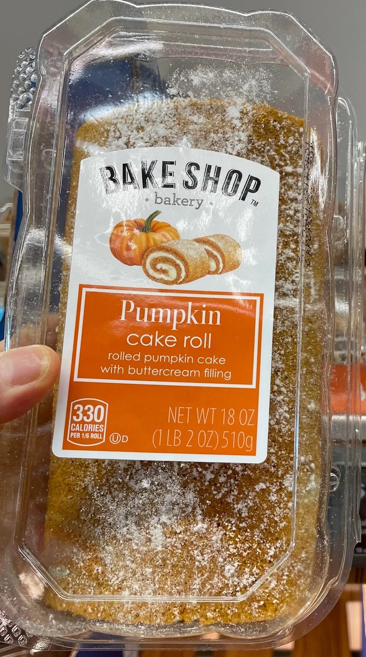 A clear plastic container holds a Bake Shop bakery pumpkin cake roll with buttercream filling. The label shows an image of rolled cake slices and lists the net weight as 18 oz and 330 calories per roll.