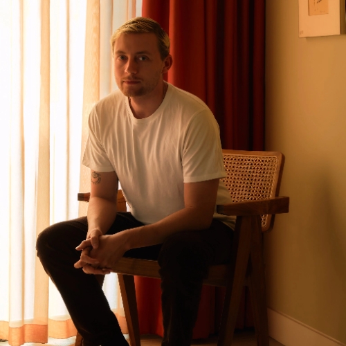 Image of a man in a white T-shirt and black pants sitting in a rattan chair in a yellow room with red curtains.