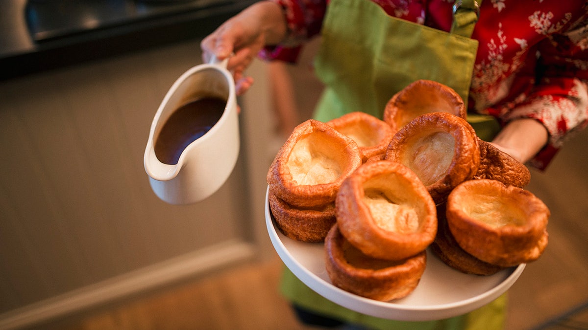 Woman in a Christmas outfit and apron carrying a plate of Yorkshire pudding puffs and a carafe of gravy.