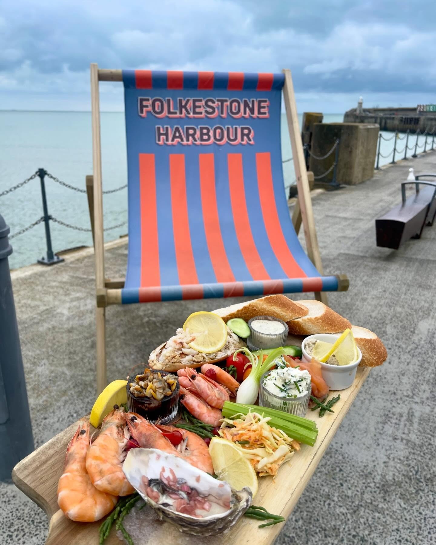 A wooden serving board with seafood and bread in front of a deckchair that says "Folkestone Harbour".