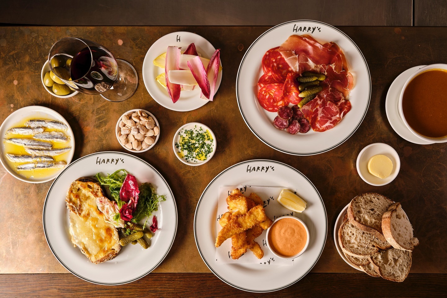 An overhead shot of several plates of food from Harry's restaurant, including cured meats, fried fish, anchovies, cheese on toast, salad, olives, nuts, and red wine.