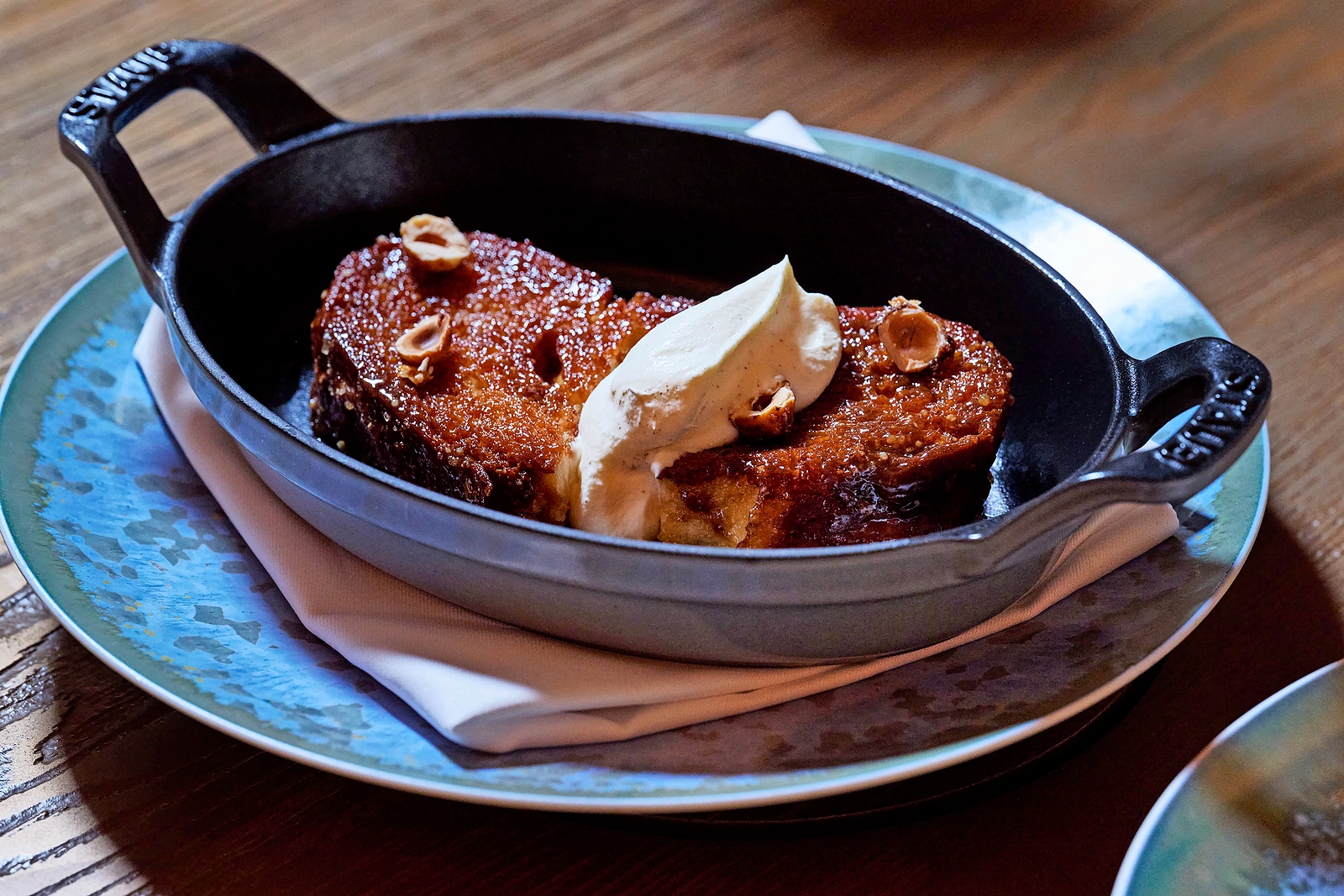 Breakfast tasting menu item from Four Seasons, a small black cast iron pan containing glazed French toast topped with whipped cream and hazelnuts, served on a blue decorative plate.