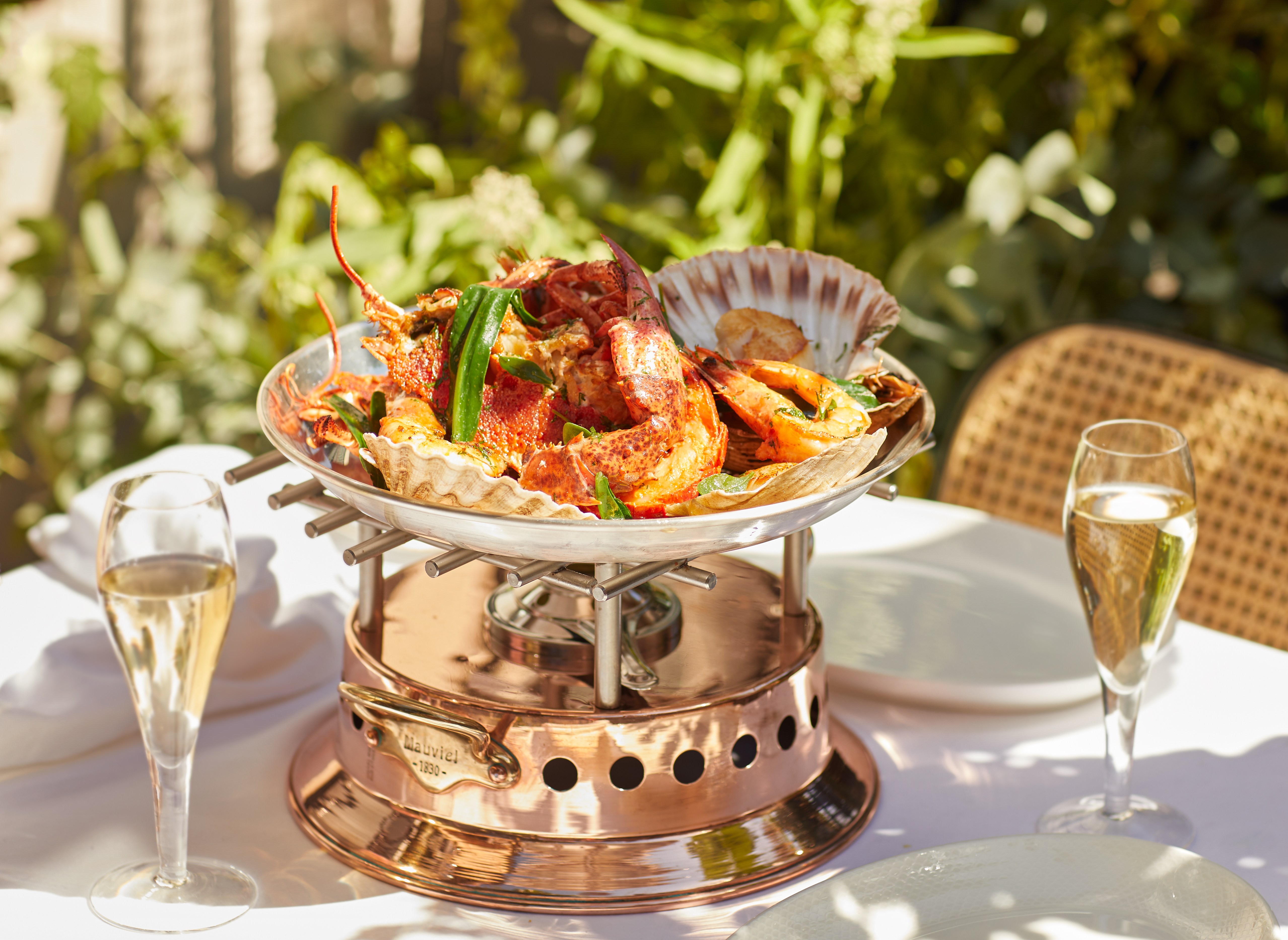 Roasted shellfish for two, including lobster, scallops, and tiger prawns with garlic butter, served in a metal platter on a copper warming stand, with two glasses of champagne on a white tablecloth.