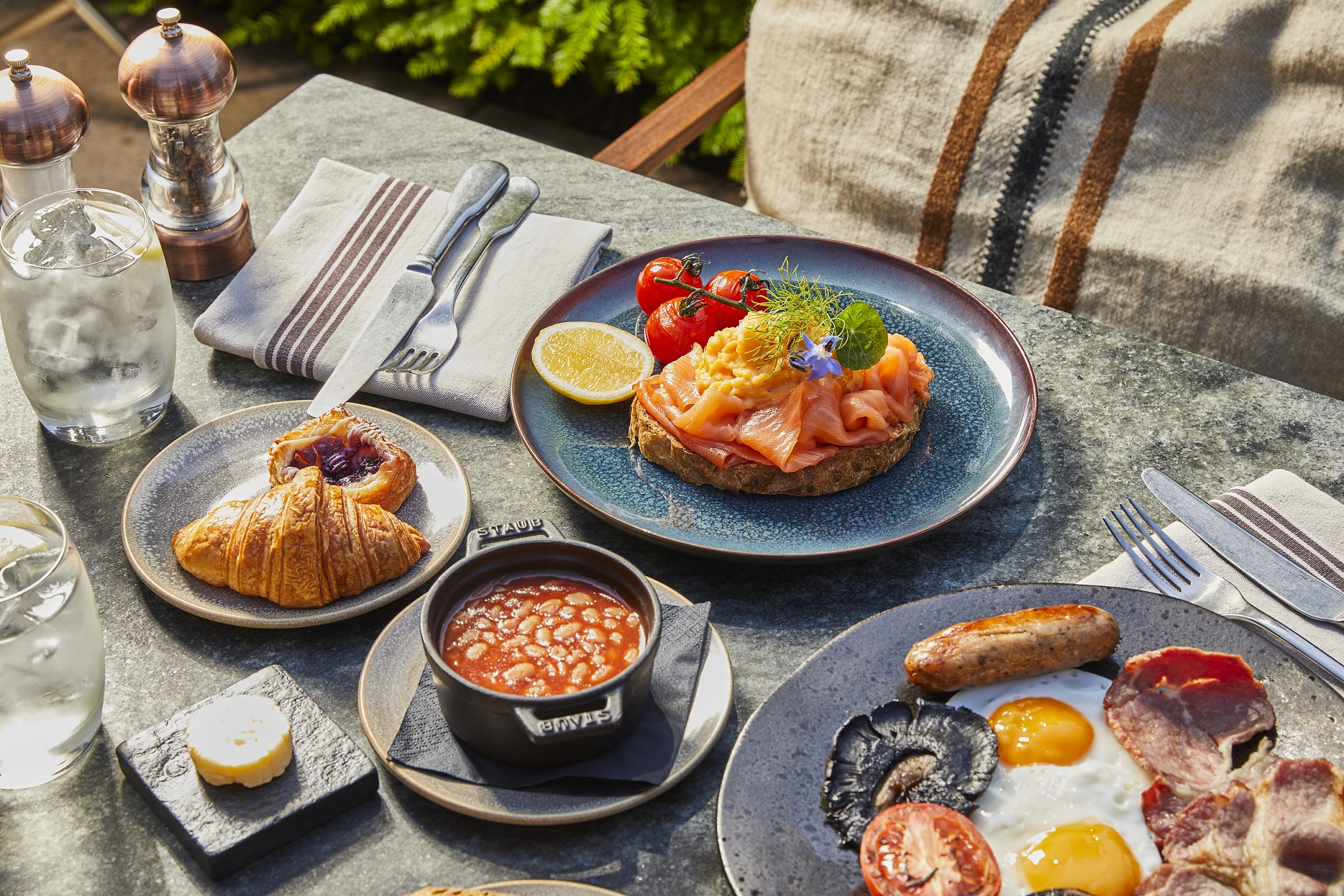 Breakfast spread with smoked salmon on toast, baked beans, croissant, and a full English breakfast with bacon, sausage, fried eggs, and mushrooms.