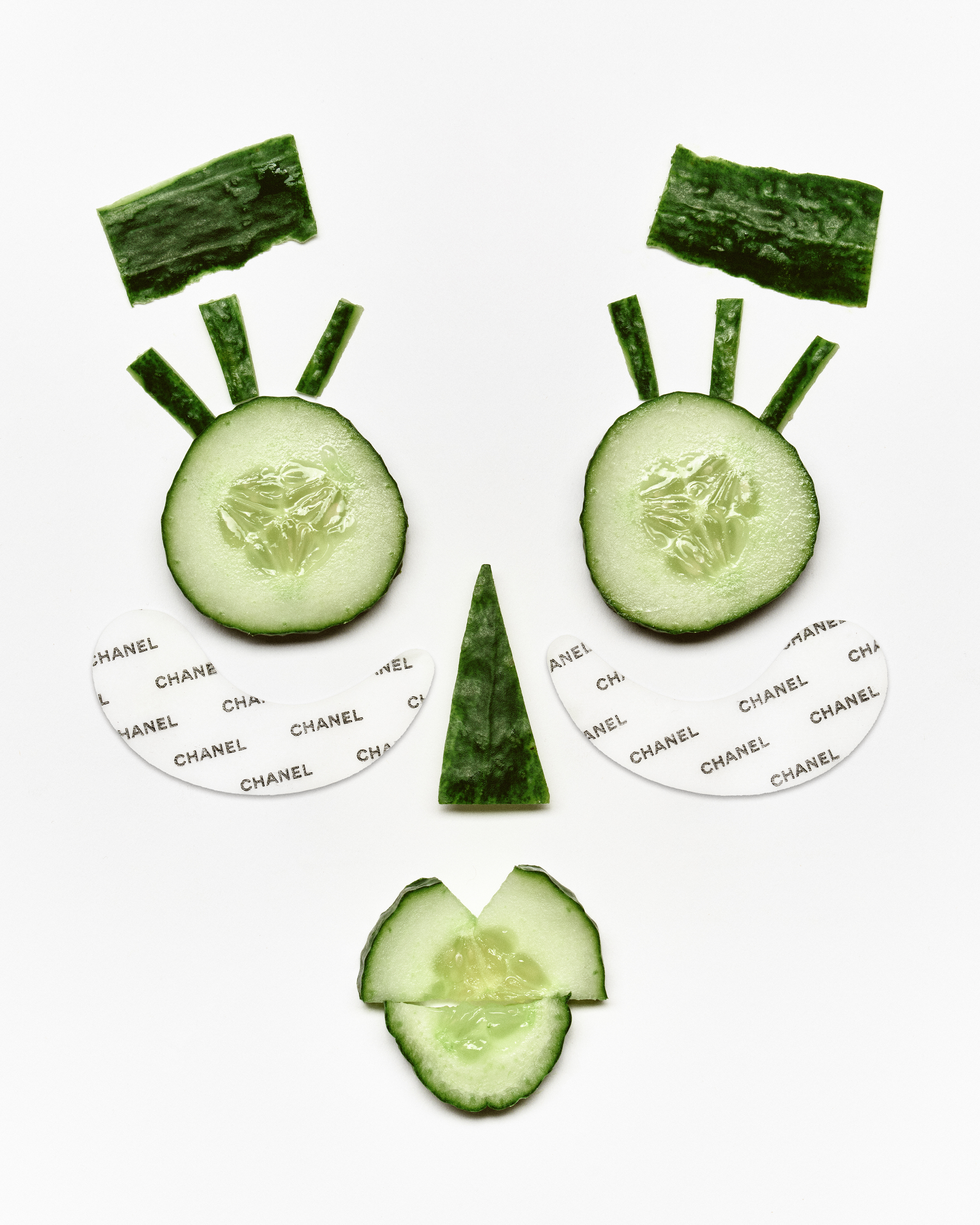 An artistic arrangement of cucumber slices and peels forming a face, with under-eye patches labeled "CHANEL."