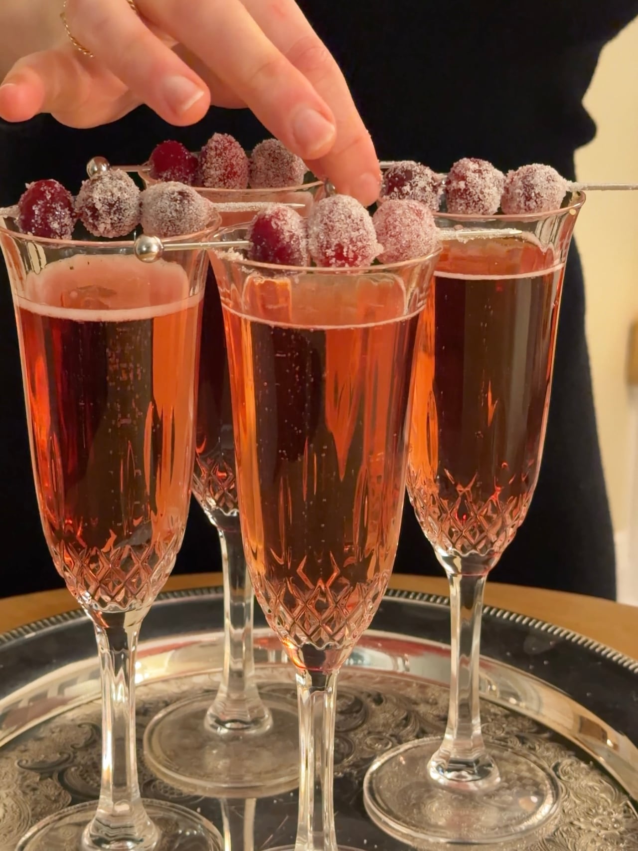 Closeup on cocktails in champagne flutes on a silver tray. They're topped with spears of sugared cranberries. 