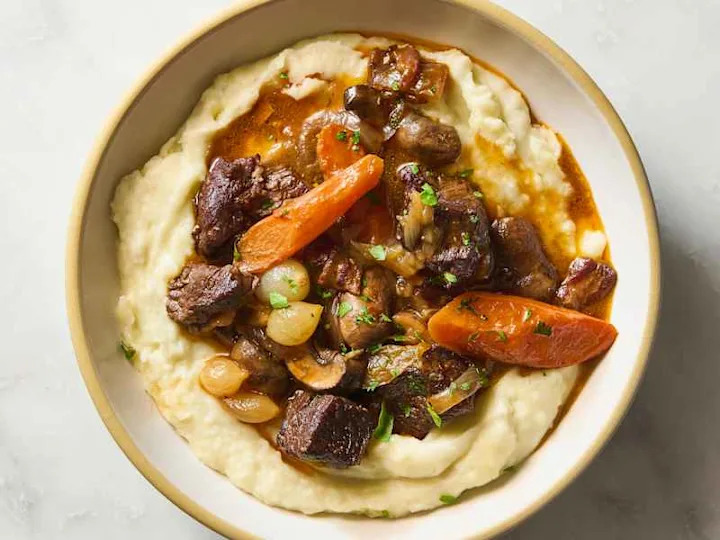 Savory beef stew with carrots, onions, and mushrooms served over creamy mashed potatoes, garnished with parsley.