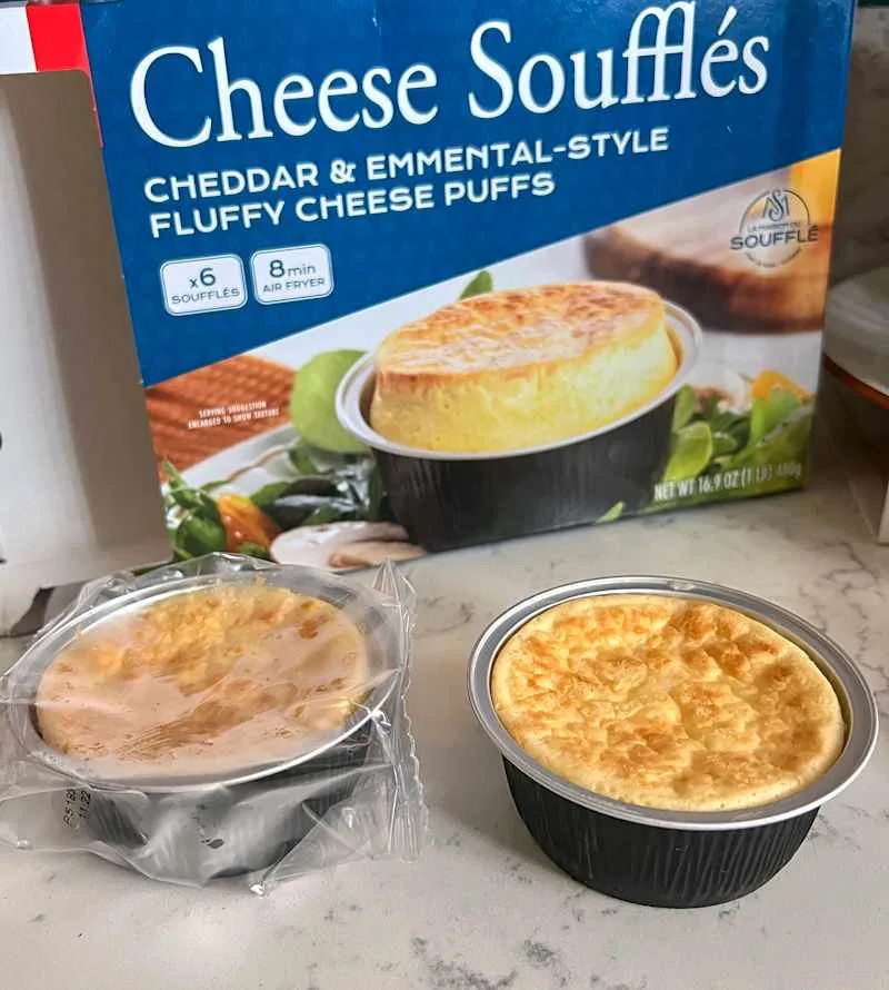 Cheese soufflés in black cups, one unwrapped and one in plastic, with a blue box in the background.