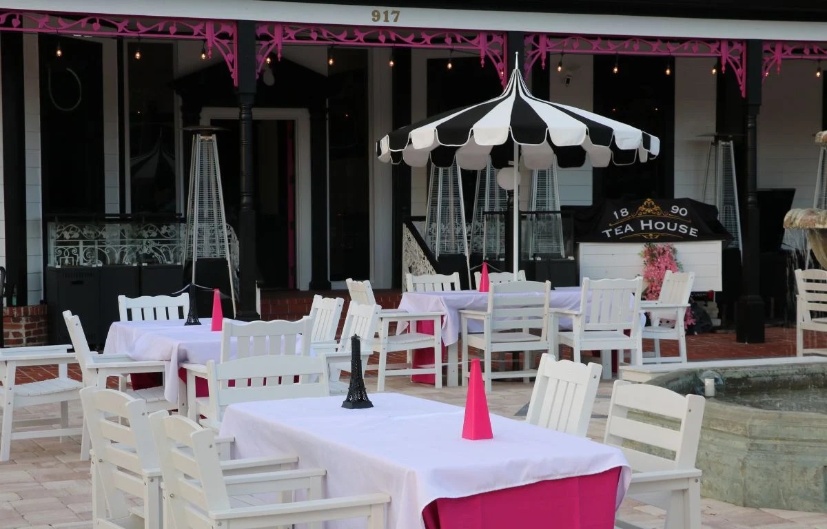 An outdoor seating patio features tables with white tablecloths and chairs scattered around the area with fans in the shape of pink pyramids and black Eiffel Towers. A black and white striped umbrella shades a table. Behind the tables is a large house painted white, black and pink. A sign in the background reads, "1890 Tea House."