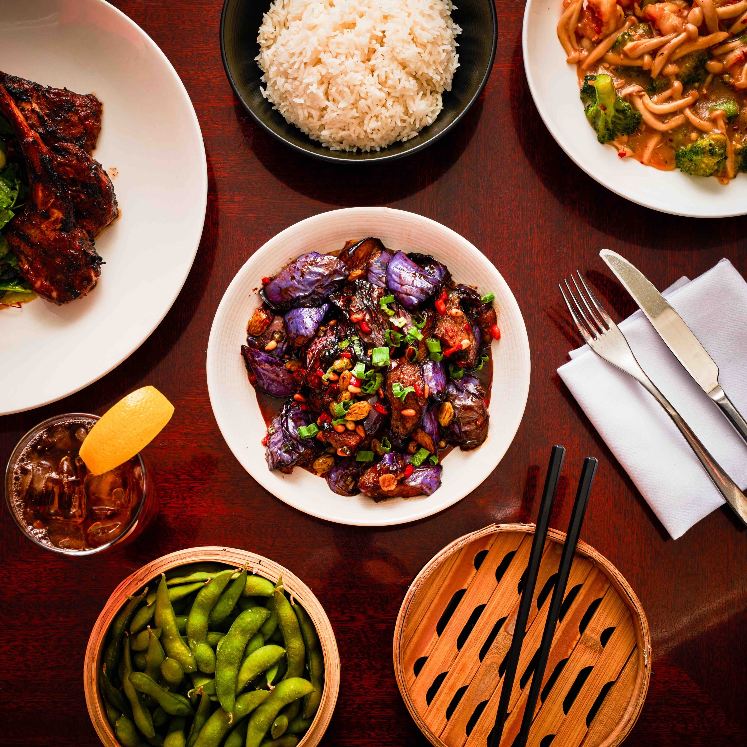 array of asian dishes including edamame, eggplant, rice, and noodles with a steam basket and a cocktail