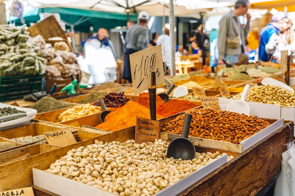 Colorful market stall showcasing spices, nuts, and food products, with people browsing in a bustling open-air environment. The scene captures a lively atmosphere of a traditional street market.