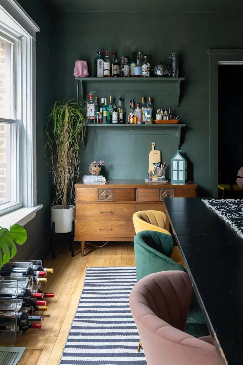Cozy dining area featuring a dark green wall, wooden sideboard, colorful velvet chairs, and a wine rack.
