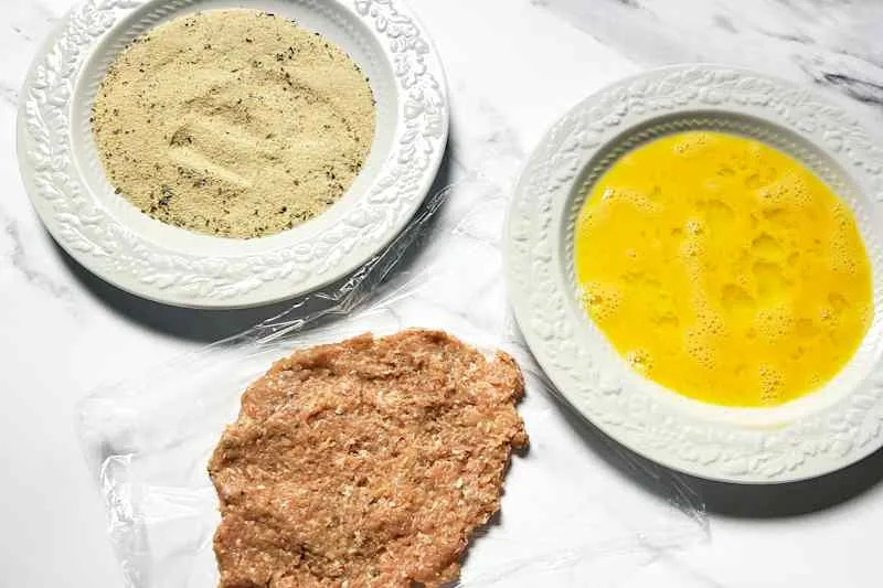 Ground meat mixture on plastic wrap, surrounded by bowls of seasoned breadcrumbs and beaten eggs.