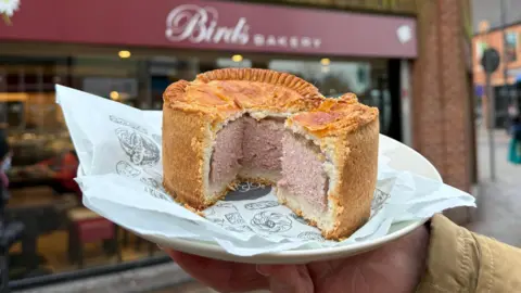 BBC A large pork pie with a quarter cut out sits on a wrapper and plate in front of a bakery. The plate is being held be someone out of shot. The bakery has a large glass front and the maroon sign reads 'Birds Bakery.' There is a pavement and road sign beside it.