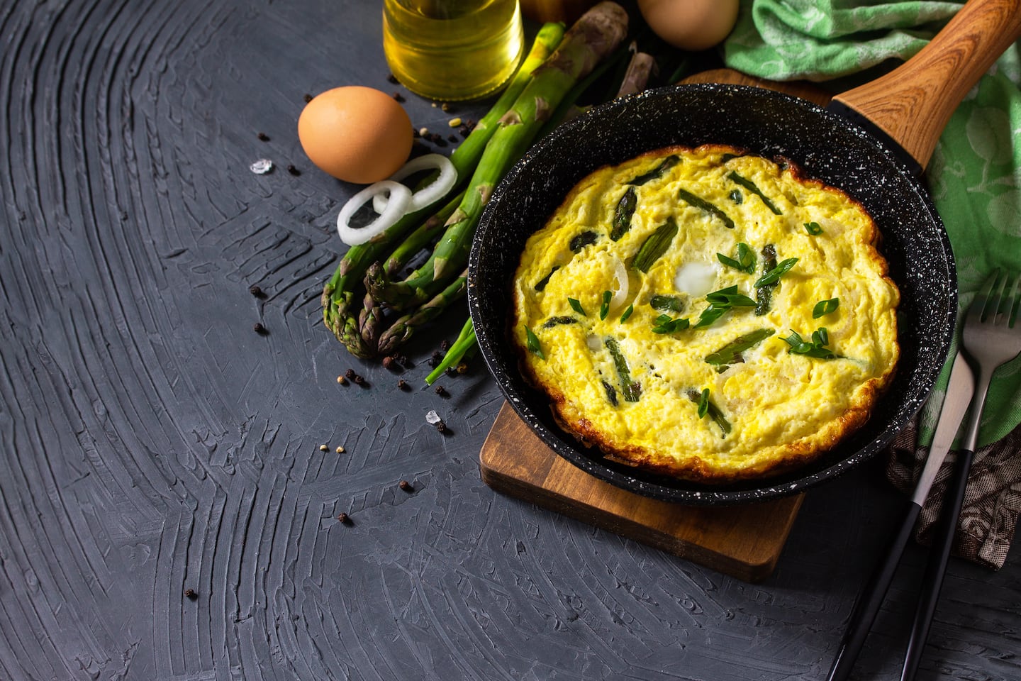 A round baking pan with a frittata with asparagus. The pan is on a wooden board, which is set on a dark surface. Beside the pan are ingredients for the frittata, including eggs and asparagus.