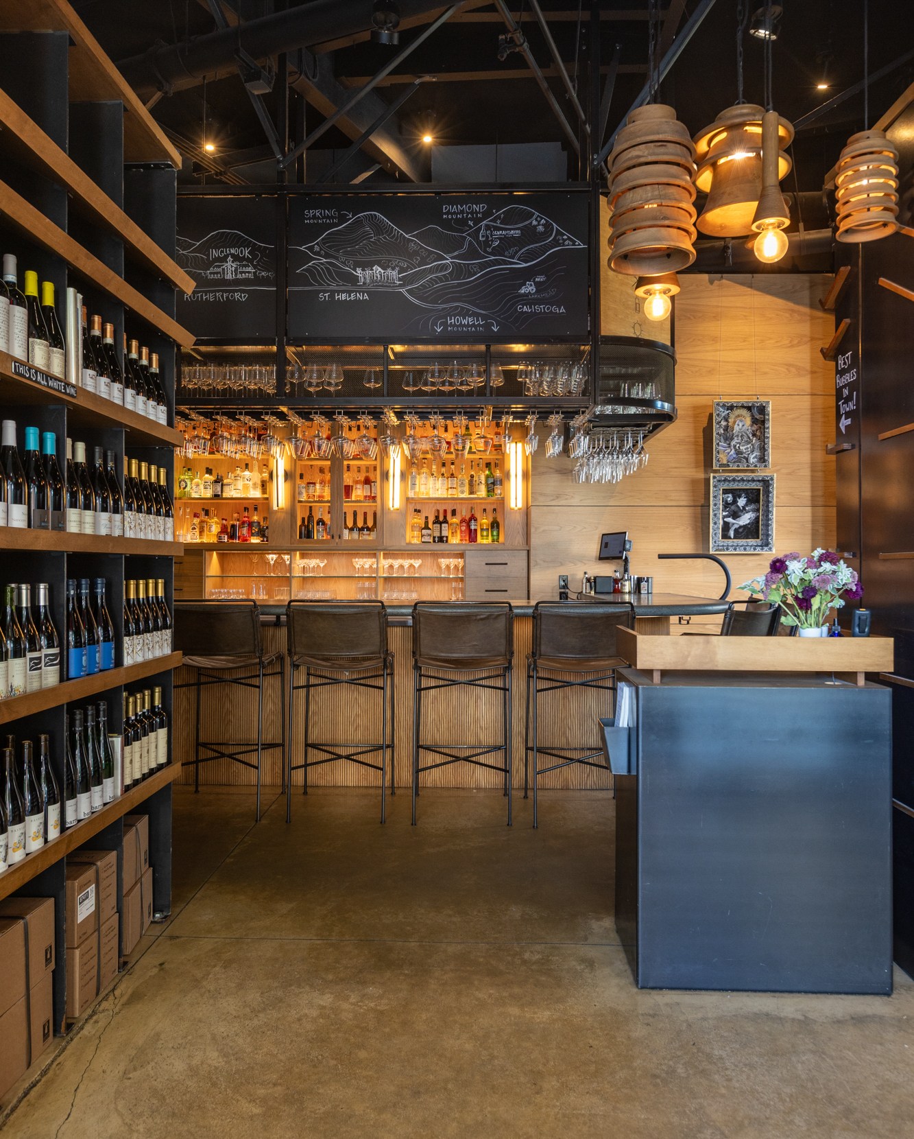 The interior of Compline in Napa, with shelves of wine bottles on the left, and a bar in the background.