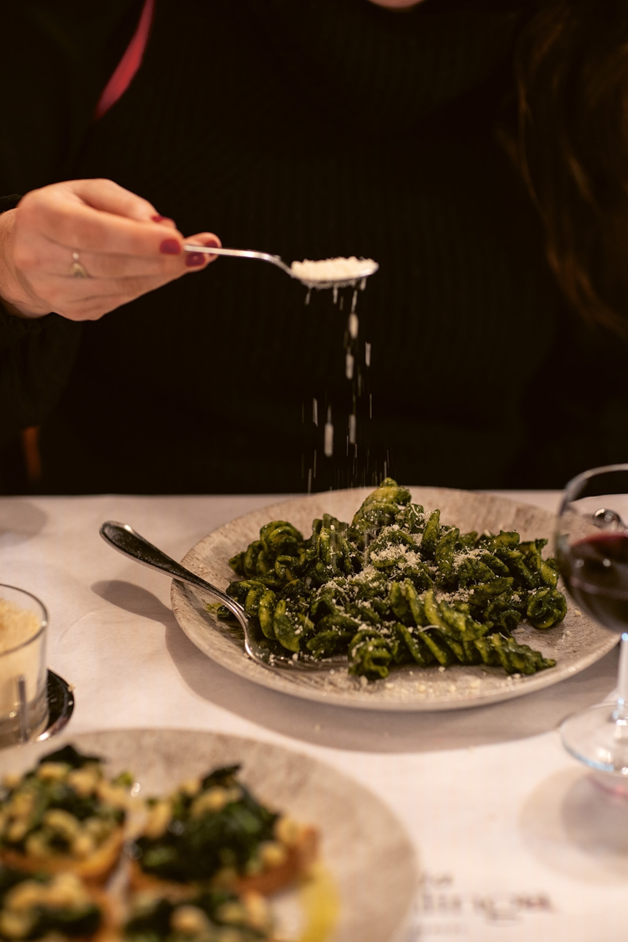 A dynamic shot of someone adding parmesan to a plate of pesto pasta at a linen-decked table.