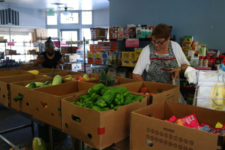 Two women sorting food items in a community food pantry with boxes of produce and shelves of packaged goods.
