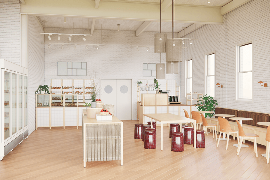 Bright, modern bakery interior with light wood flooring and white brick walls. The space features display shelves with baked goods on the left, a central wooden table with decorative items, and a seating area on the right with orange chairs and a long brown cushioned bench. Large windows on the right wall allow natural light to fill the room. Pendant lights hang from the ceiling, and plants add greenery to the space.