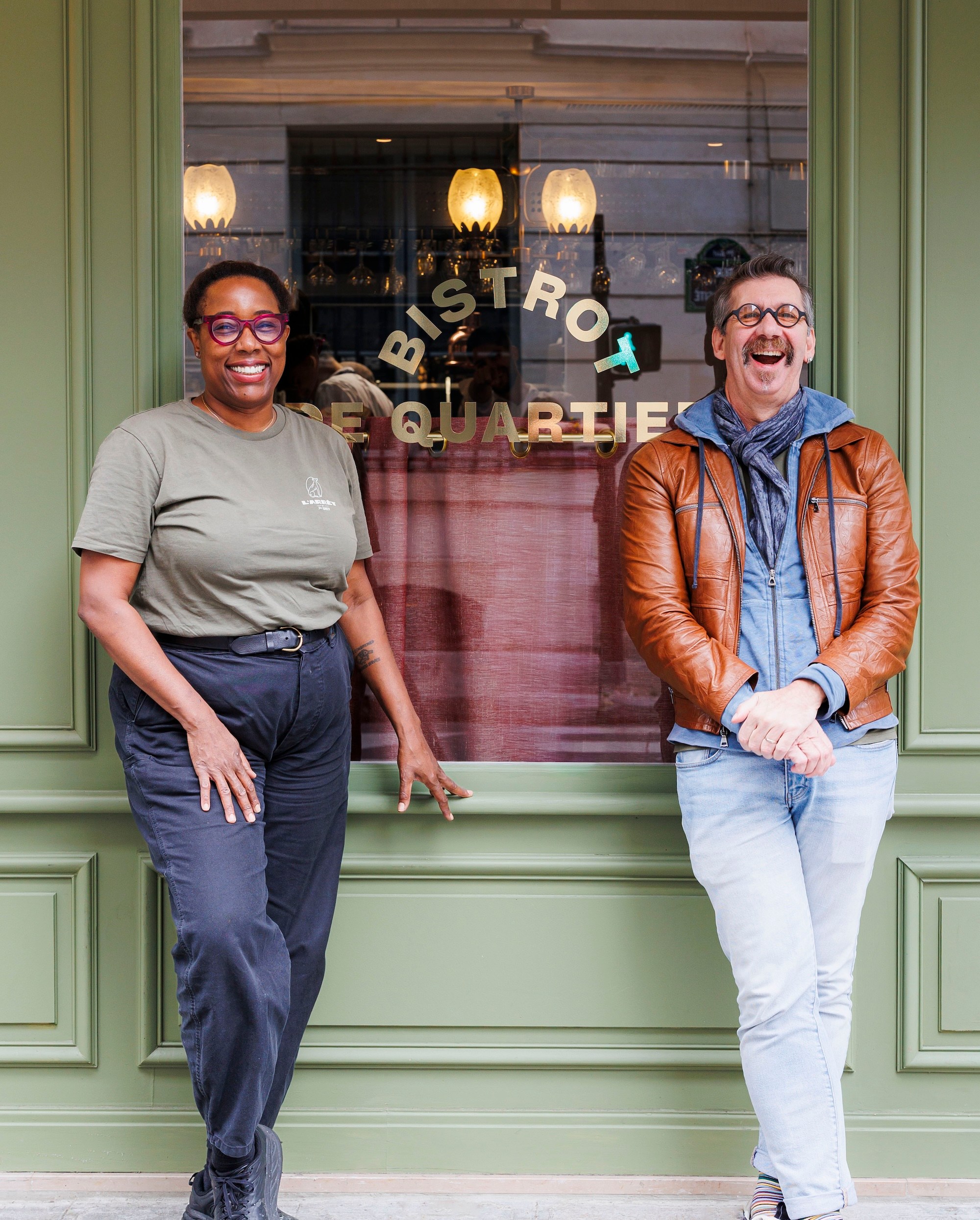 Mashama Bailey and Johno Morisano stand outside their restaurant, leaning against a window with the word “bistrot” visible.