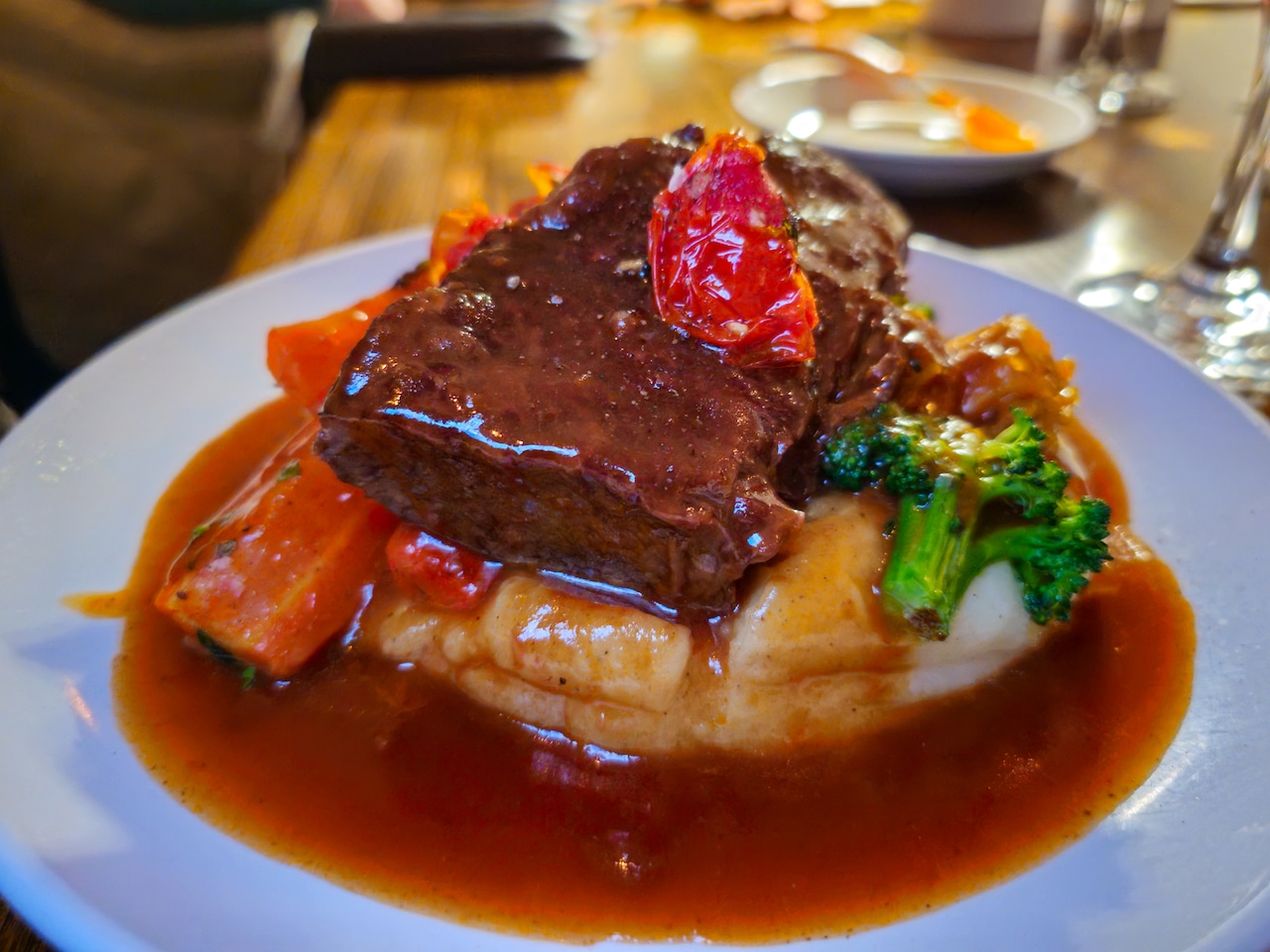 Beef short rib on a bed of mashed potatoes, along with roasted vegetables, all covered in sauce.