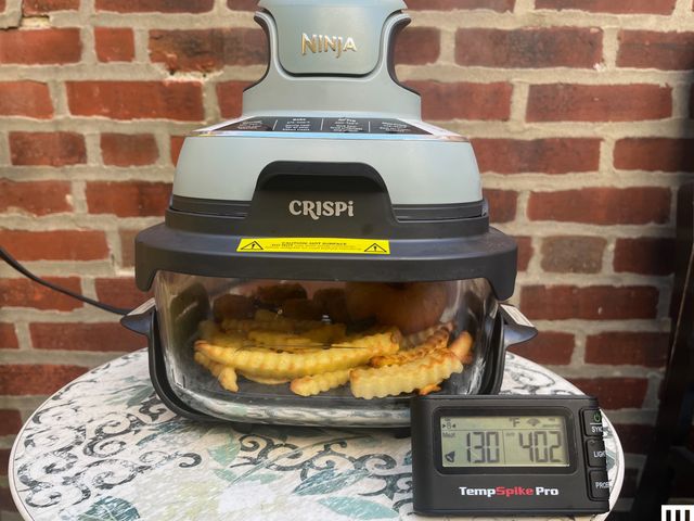 Closeup of the Ninja Crispi, a portable airfryer comprised of a clear dish on the bottom and a two-tier funnel-shaped device attached on top, sitting on a small table outdoors with a digital thermometer in front