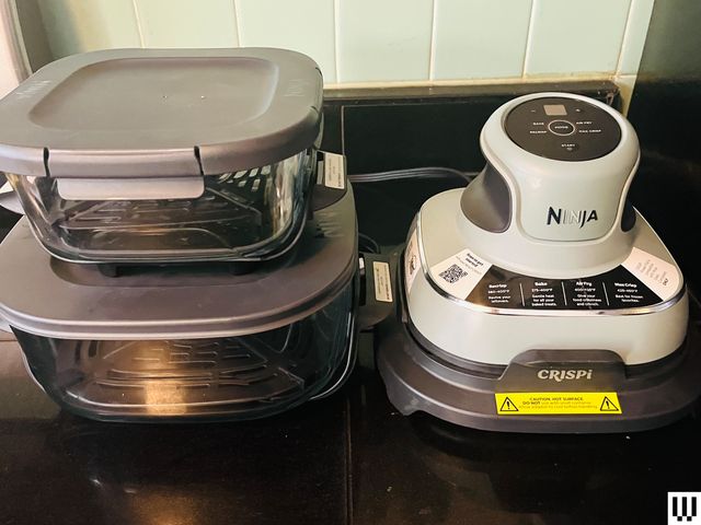 Closeup of the Ninja Crispi portable air-fryer system, including 2 stacked clear dishes on the left and a and a two-tier funnel-shaped device on the right, sitting on a stovetop