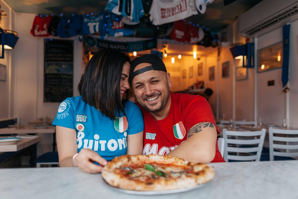 This photo shows Austria and Cirio Iovine smiling and leaning their heads together, with a Margherita pizza in front of them, as they sit in the dining area of Song E' Napule