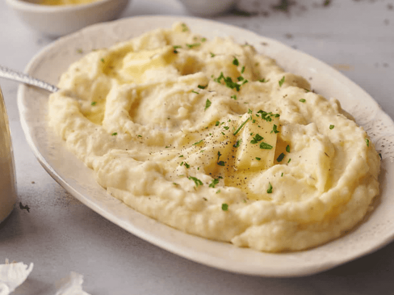 A plate of mashed potatoes topped with melted butter, ground black pepper, and chopped parsley, served with a spoon.