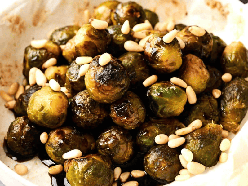 A plate of roasted Brussels sprouts topped with pine nuts on parchment paper.