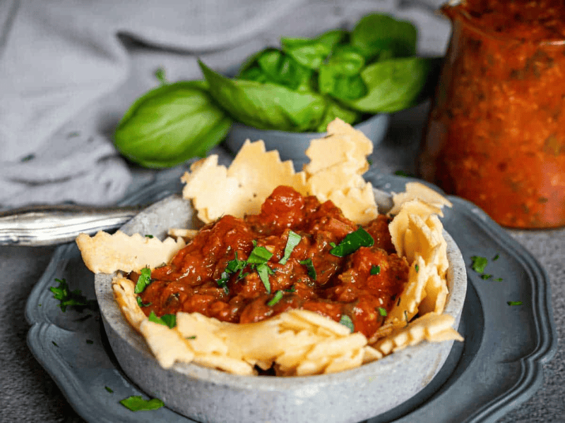A bowl filled with tomato-based pasta sauce, garnished with herbs, surrounded by pieces of broken parmesan cheese, with fresh basil and sauce jar in the background.