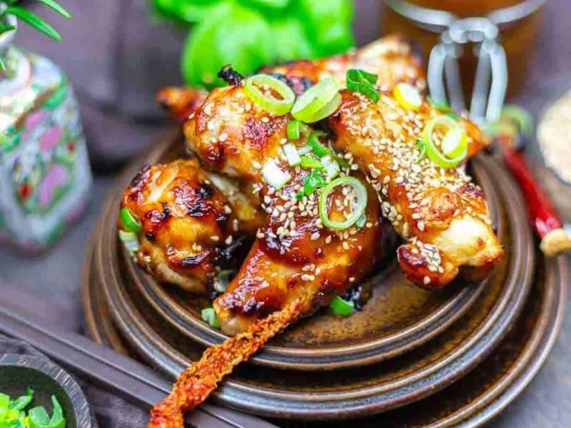 Oven baked BBQ wings garnished with sesame seeds and sliced green onions, served on a brown ceramic plate with decorative items in the background.