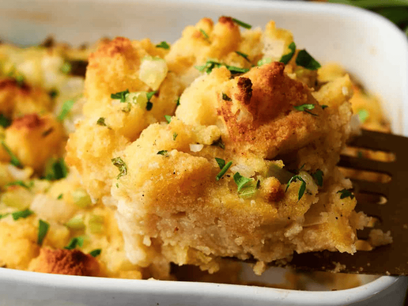 A metal spatula lifts a serving of baked stuffing topped with chopped parsley from a white casserole dish.