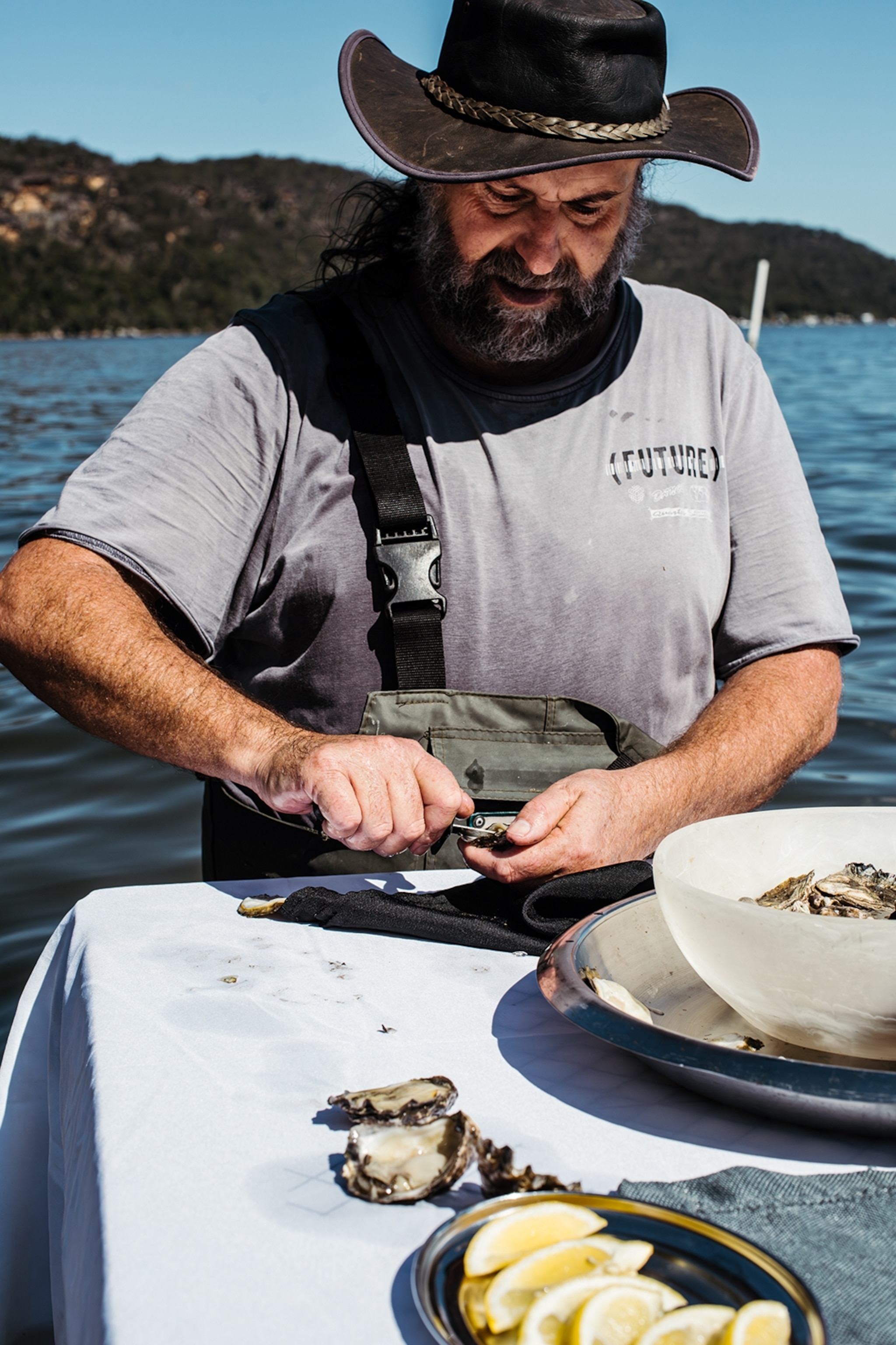 A friendly-looking older man with a leather hat, shucking oysters at the edge of a half-submerged table in the ocean.
