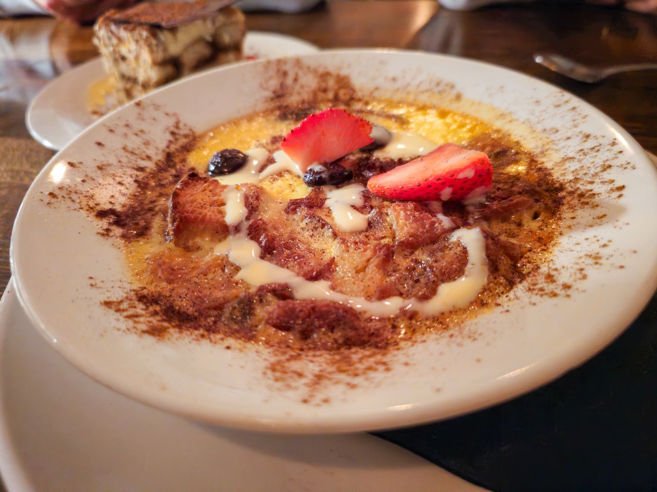 A shallow bowl of bread pudding, garnished with cocoa powder and fresh fruit.