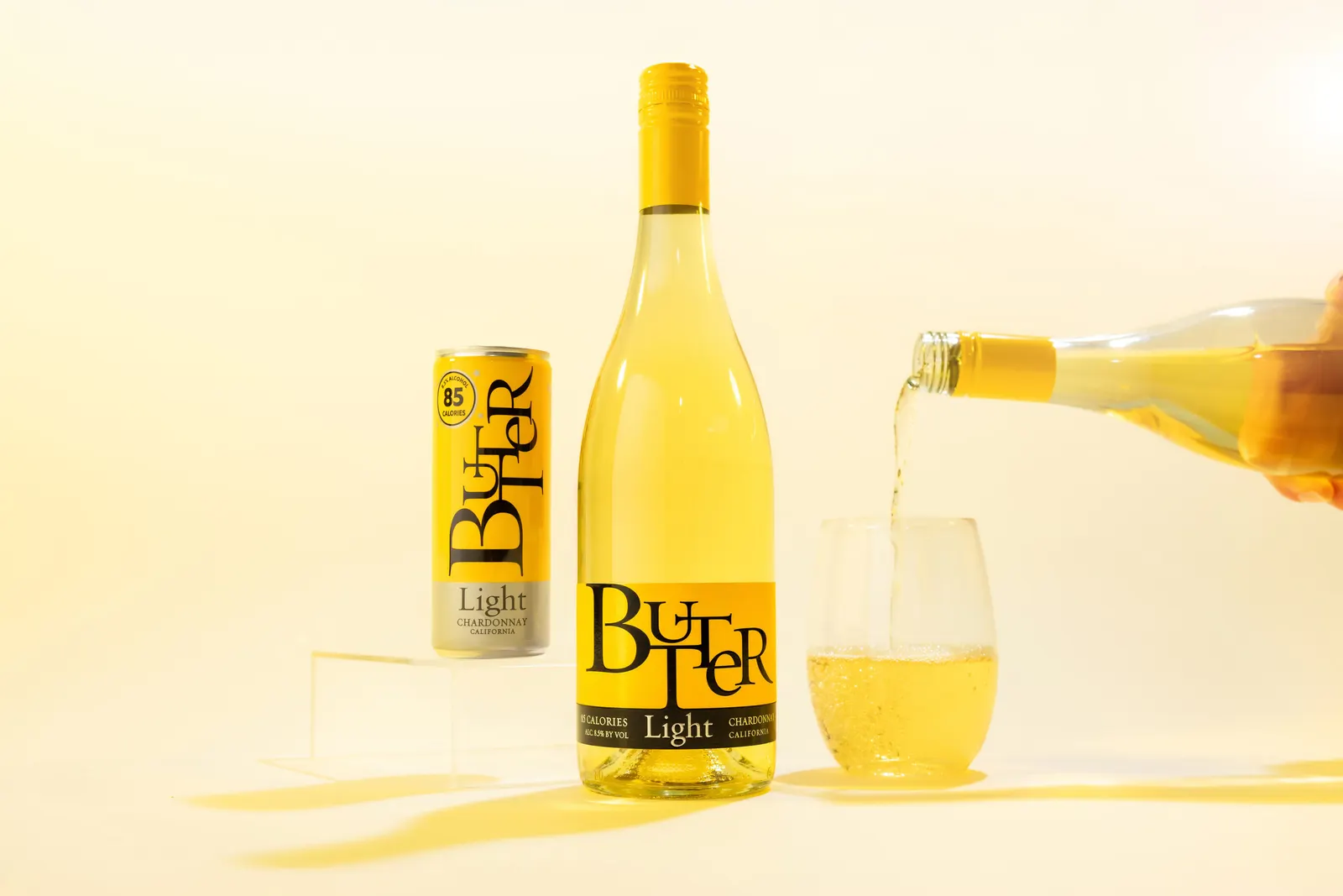 A person pours wine from a bottle into a glass that is next to a bottle and a can of ButterLight chardonnay.