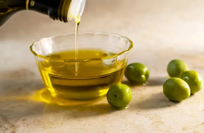 Olive oil being poured into a glass bowl, with several green olives nearby on a marble surface