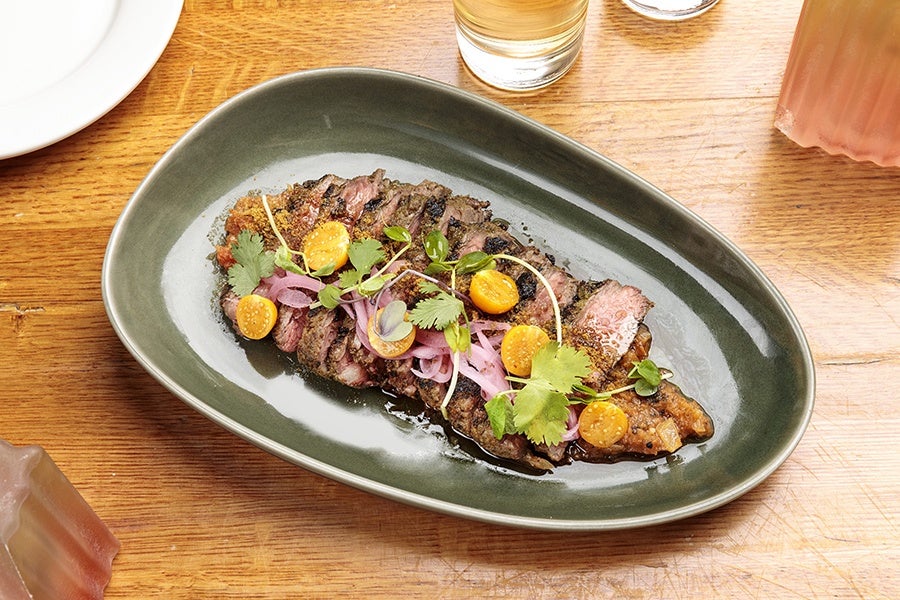Sliced grilled steak served on a dark green oval plate, topped with pickled red onions, halved yellow cherry tomatoes, fresh cilantro leaves, and microgreens, with a light sauce underneath. The plate is on a wooden table with a glass of water and a pink drink nearby.