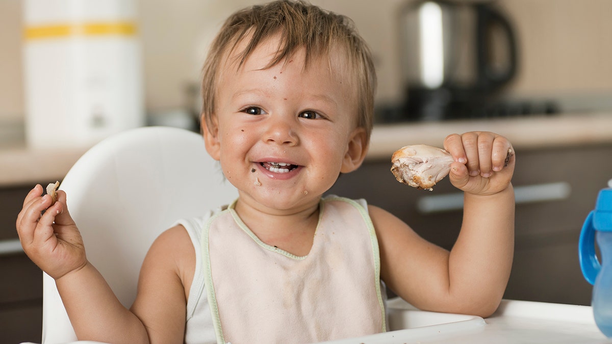 Baby boy eating chicken leg, smiling in high chair in kitchen