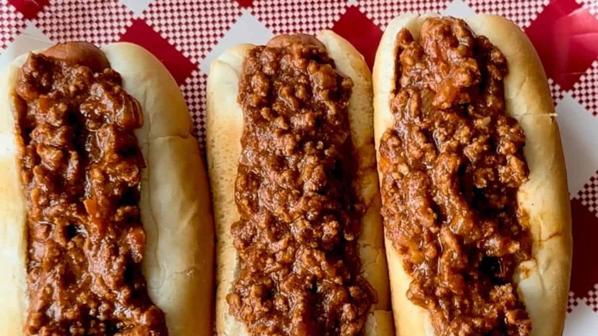 Hot dogs topped with meat chili lined up on red and white tablecloth.