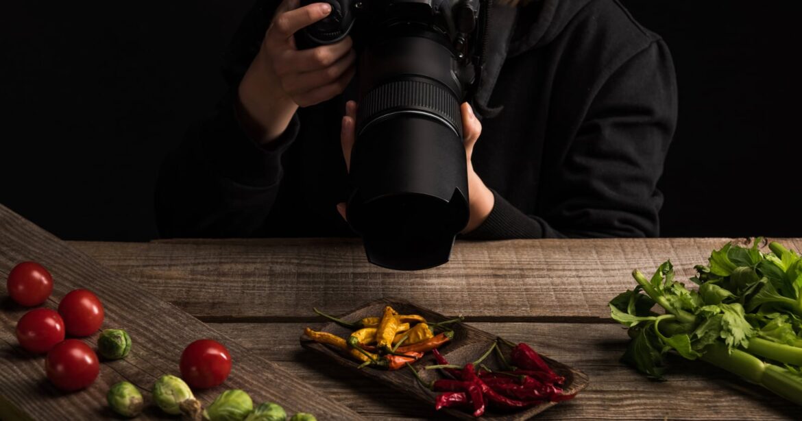 A person holding a camera photographs assorted vegetables, including cherry tomatoes, Brussels sprouts, celery, and colorful chili peppers, arranged on a rustic wooden table against a dark background.