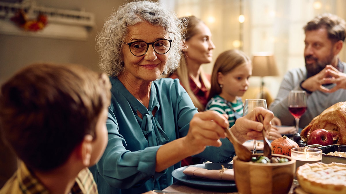 Family eating holiday meal around the table, grandma seen serving food, looking at young grandson.