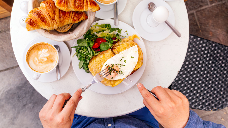 Person cutting into a croque madame at a table with coffee and croissants.