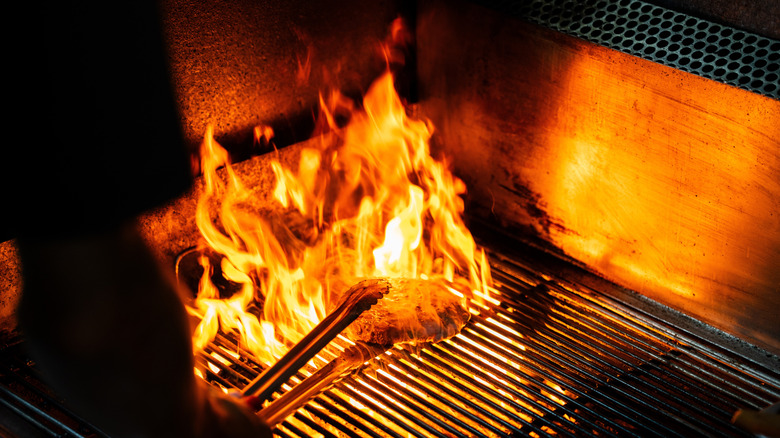 Someone flipping meat with tongs on top of a grill with large, glowing flames